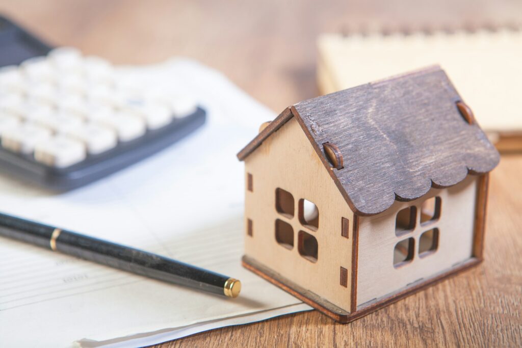 Miniatura de casa ao lado de calculadora e caneta sobre documentos, representando o cálculo do imposto sobre ganho de capital na venda de imóvel